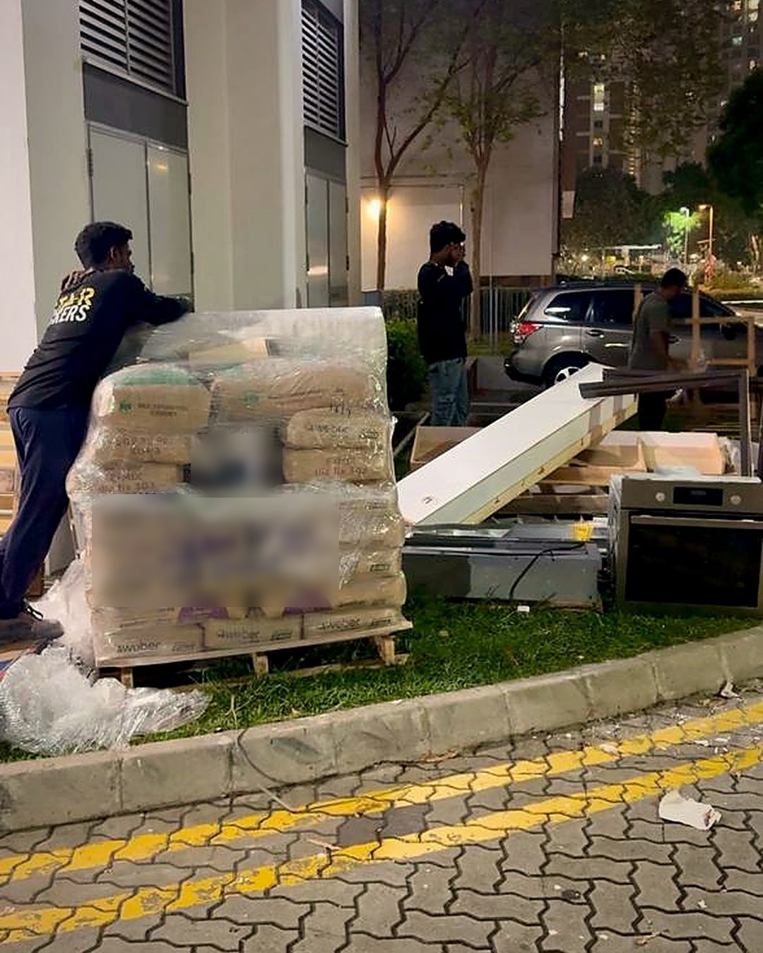 A wrapped pallet of renovation materials at the foot of an HDB block at night, a mover beside it