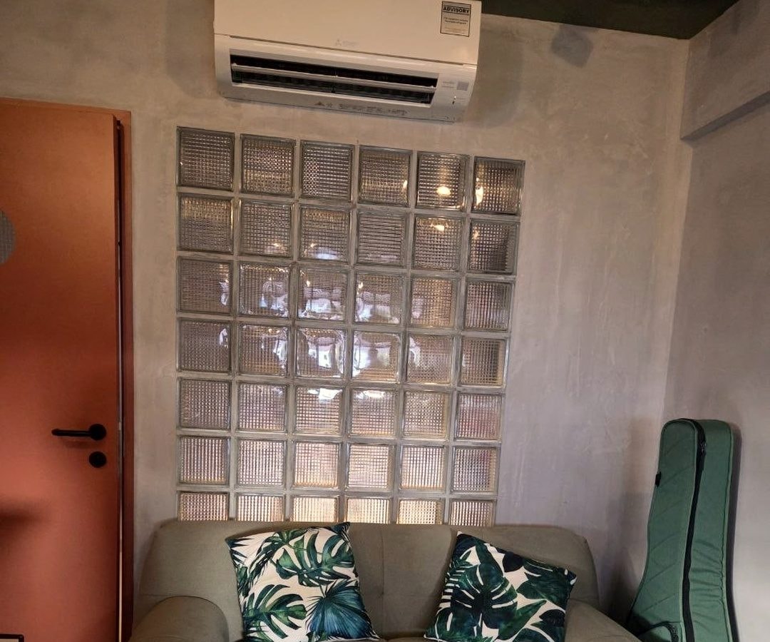 A compact HDB living room with a wall-mounted aircon above a glass block wall and a sage sofa