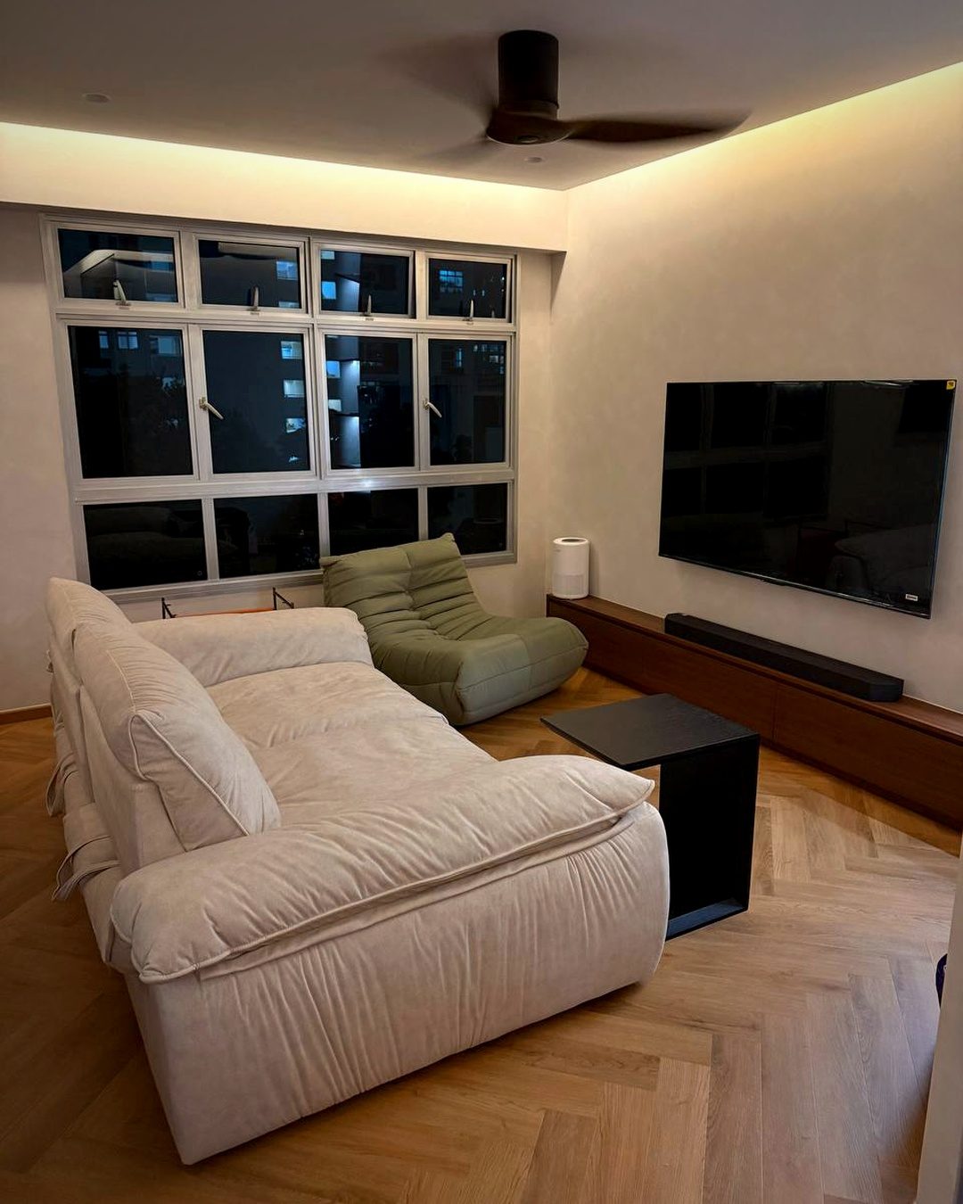 A living room with a lit ceiling cove, a ceiling fan, large windows and a wall-mounted TV